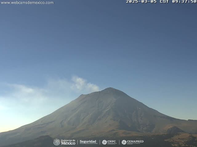 A large, gray volcano with a plume of smoke emanating from its peak is visible under a clear blue sky.