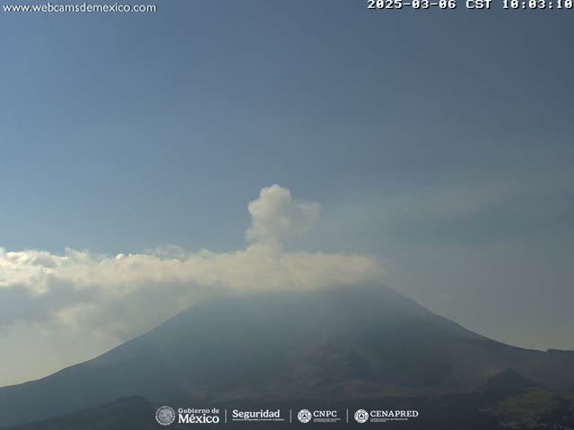 A large volcano is seen under a mostly clear sky with some clouds, and a small plume of smoke or ash is emanating from its peak.