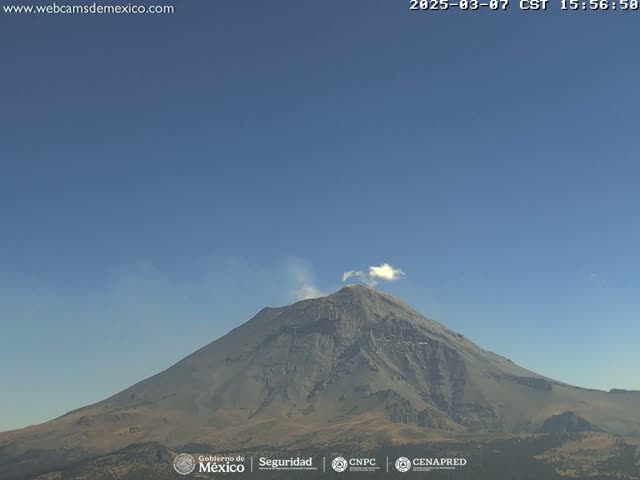 A large, gray volcano with a small plume of smoke erupting from its summit is visible under a clear blue sky.