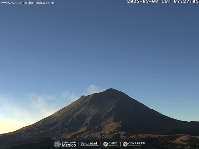 A large, dark-colored volcano is seen under a clear, light-blue sky with a small plume of smoke emanating from its peak.