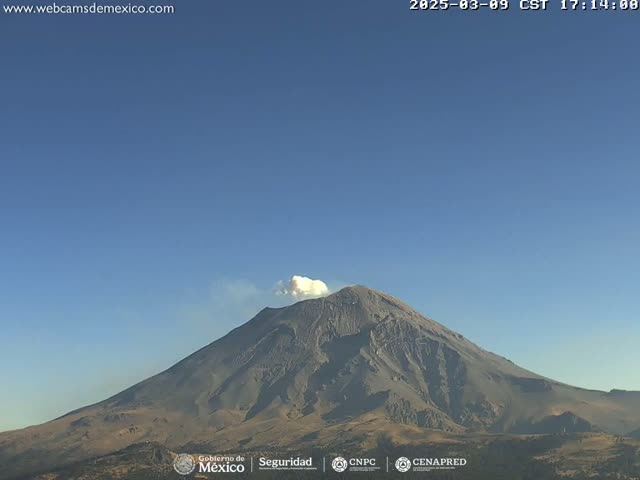 A large, dark-grey volcano with a plume of white smoke erupting from its peak is visible under a clear blue sky.