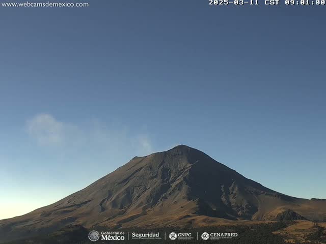 A large, dark-colored volcano with a plume of light-colored smoke rising from its peak is visible under a clear blue sky.