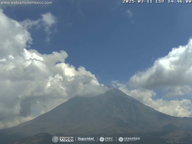 A large, snow-capped mountain is partially obscured by fluffy white clouds under a mostly clear blue sky.