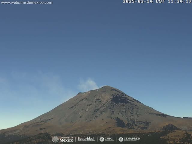 A large, dark-grey volcano with a plume of light-grey smoke emitting from its peak is visible under a clear, light-blue sky.