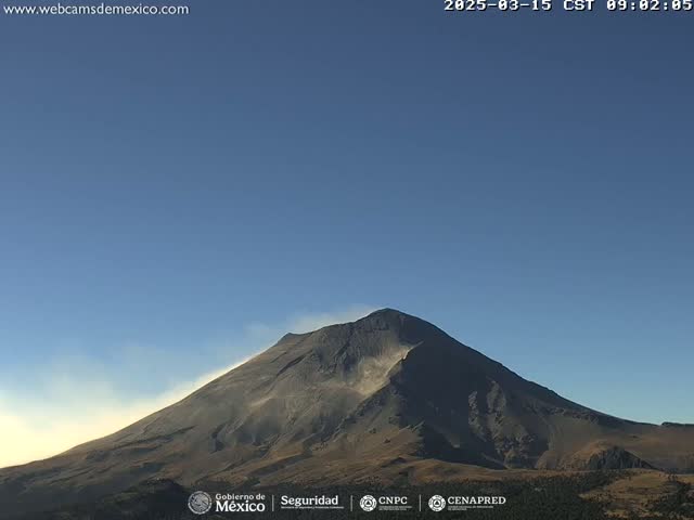 A large, dark-colored mountain with a light-colored plume of ash or dust emanating from its side is visible under a clear blue sky.