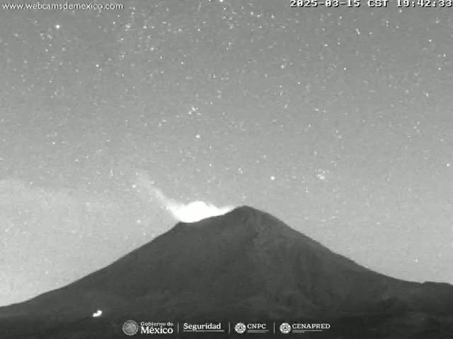 A nighttime, grayscale image shows a volcano emitting a plume of smoke or ash under a starry sky.