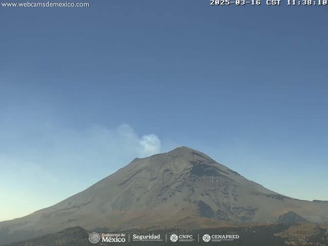 A large, dark-grey volcano emits a small plume of light-grey smoke under a clear, light-blue sky.