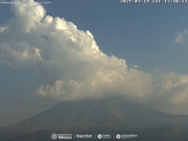 A large, fluffy cumulus cloud formation partially obscures a hazy mountain range under a mostly clear, light-blue sky.