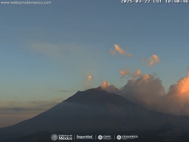 A large, dark mountain is partially obscured by clouds illuminated by the setting sun in a mostly clear sky.