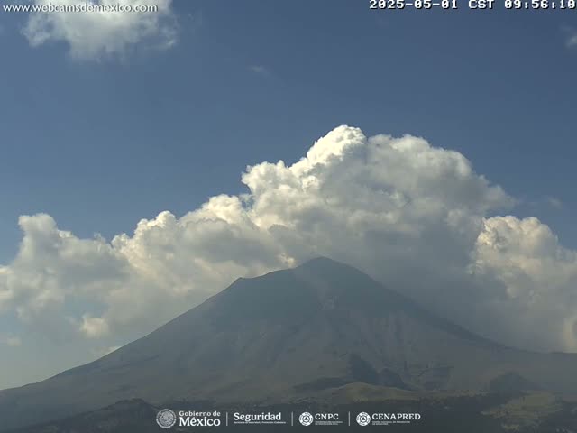 A large, dark-grey volcano is partially obscured by fluffy white cumulus clouds under a mostly clear blue sky.