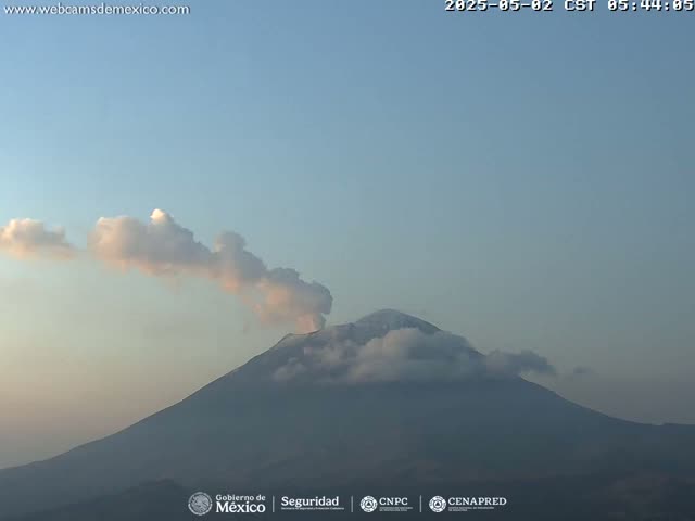 A snow-capped volcano erupts a plume of light-pinkish smoke under a clear, pale-blue sky.