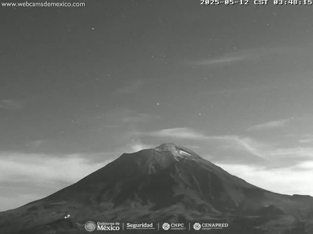 A grayscale nighttime image shows a large, snow-capped volcano under a slightly cloudy sky dotted with stars.