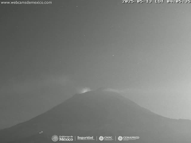 A nighttime, low-light image shows a volcano emitting a faint plume of smoke under a mostly clear sky.