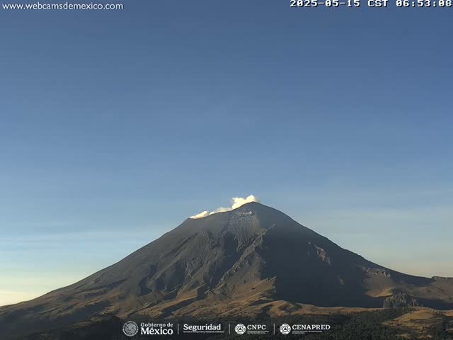 A large, dark-colored volcano with a small plume of smoke erupting from its peak is visible under a clear blue sky.
