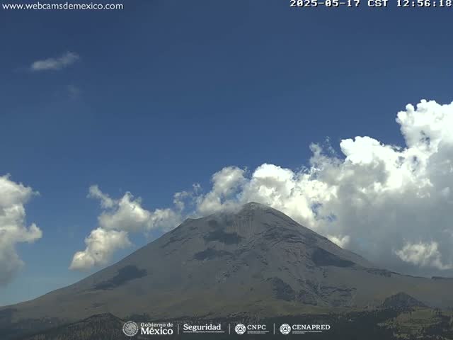 A large, gray volcano is visible under a mostly clear blue sky with scattered cumulus clouds.