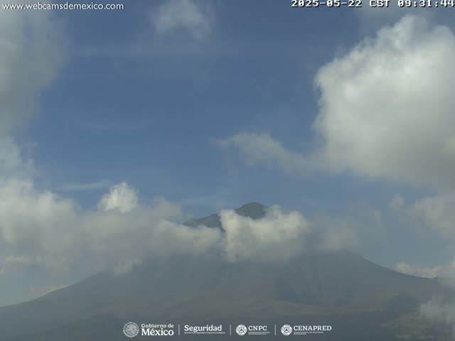 A mostly clear, blue sky with scattered clouds partially obscures a large, dark mountain.