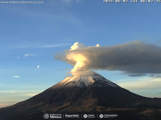 A snow-capped volcano erupts a plume of ash and steam into a mostly clear blue sky.