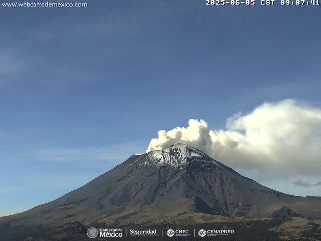 A snow-capped volcano erupts a plume of smoke under a clear blue sky.