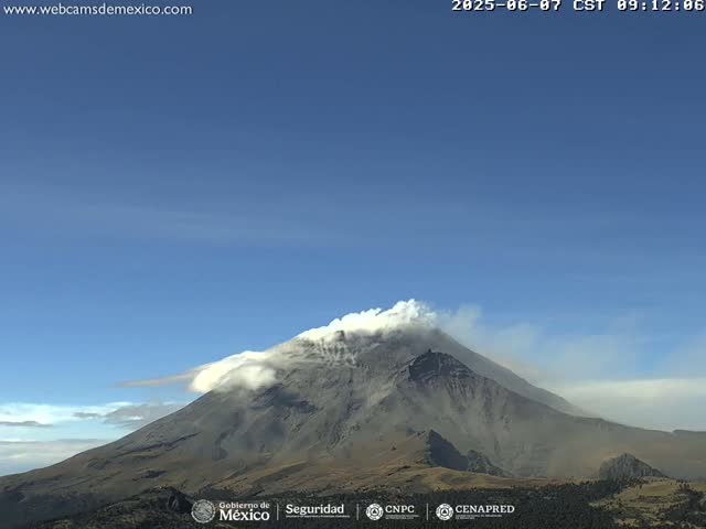 A large, dark-grey volcano with a plume of white smoke billowing from its peak is visible under a clear blue sky.