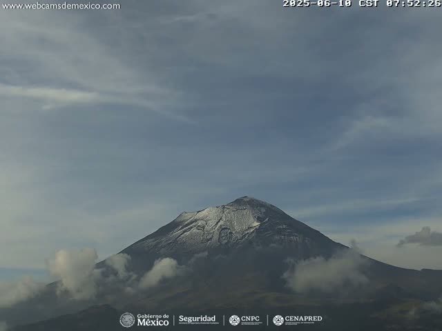 A snow-dusted, dark mountain is partially obscured by clouds under a mostly cloudy sky.
