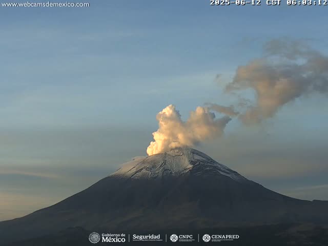 A snow-capped volcano erupts a plume of light-colored smoke under a clear, twilight sky.