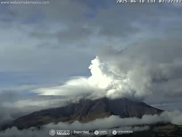 A large volcano is partially obscured by a plume of white smoke and low-lying clouds under an overcast sky.