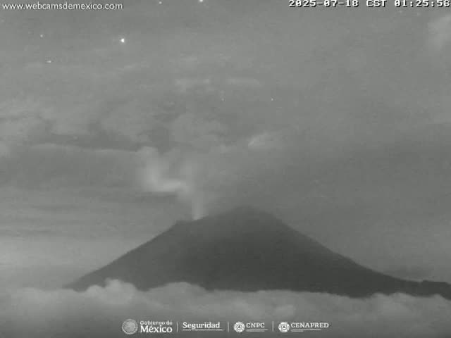 A nighttime, low-light image shows a volcano emitting a plume of smoke or ash under a cloudy sky.