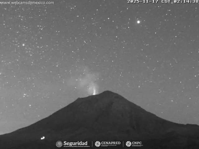 A grayscale night vision image displays a dark volcano actively emitting a bright plume of gas under a clear, starry night sky.
