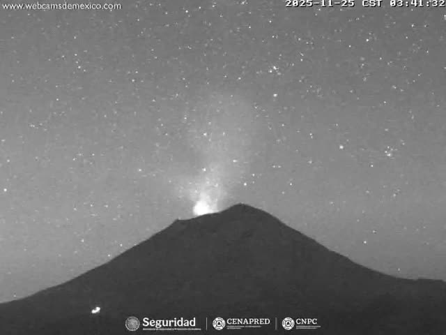 A grayscale image captures a volcano erupting with a bright plume from its summit against a clear, star-filled night sky.