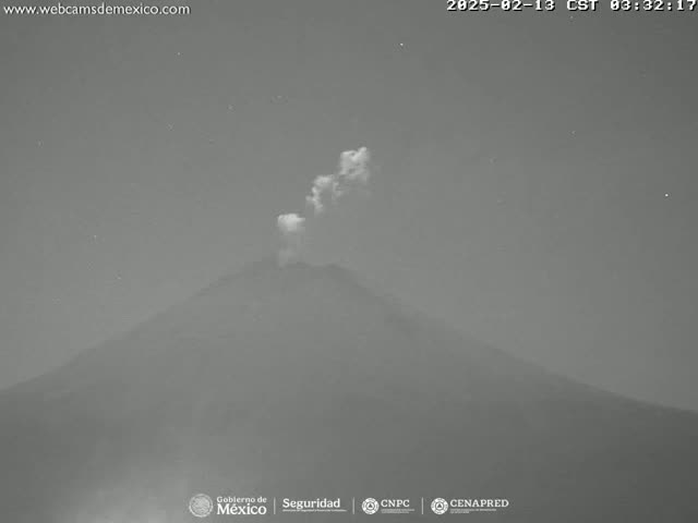 A nighttime, low-light image shows a volcano emitting a small plume of smoke or ash under a clear, starry sky.