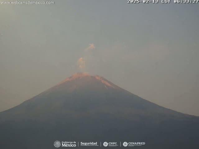 A large volcano with a plume of smoke emitting from its peak is visible under a hazy sky.