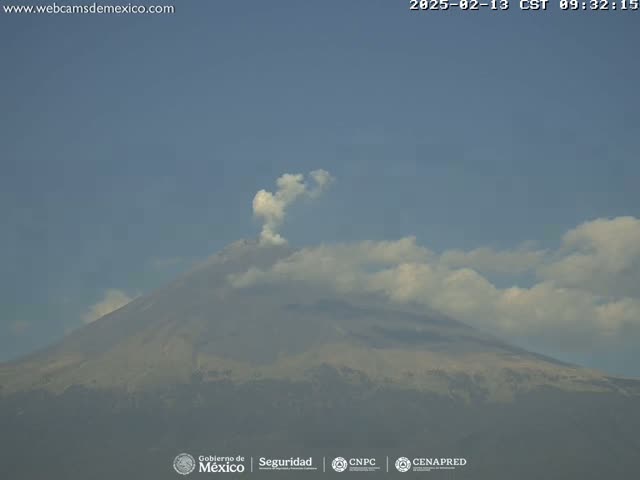 A large volcano with a light plume of smoke is visible under a mostly clear sky with scattered clouds.