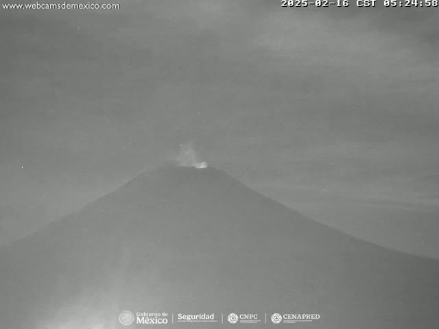 A dark, overcast nighttime image shows a volcano emitting a faint plume of smoke or ash.