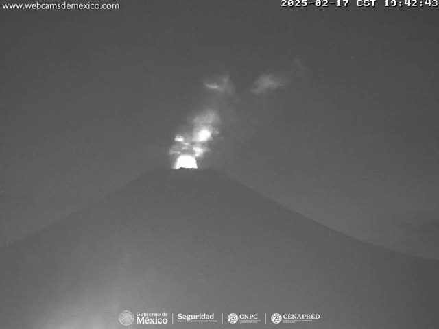 A nighttime, low-light image shows a volcano emitting a plume of light-colored smoke or ash under a mostly clear sky.