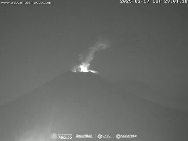 A nighttime, grayscale image shows a volcano erupting with a plume of smoke and glowing lava visible against a dark sky.
