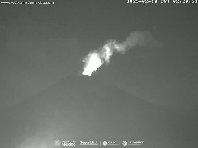 A nighttime, low-light image shows a volcano erupting with a bright plume of smoke and ash rising into the dark sky.