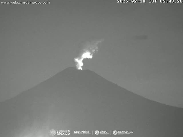 A nighttime, grayscale image shows a volcano erupting with a bright plume of smoke and ash against a dark sky.