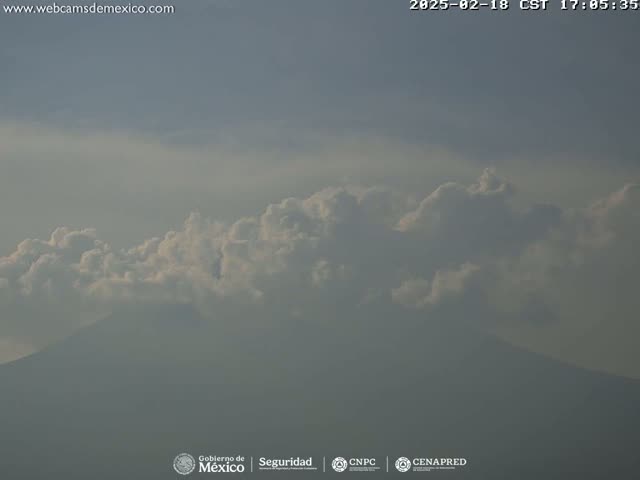 A hazy mountain is partially obscured by a layer of fluffy white clouds under a mostly clear sky.