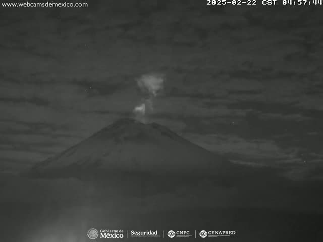 A nighttime, low-light image shows a volcano emitting a plume of smoke under a cloudy sky.