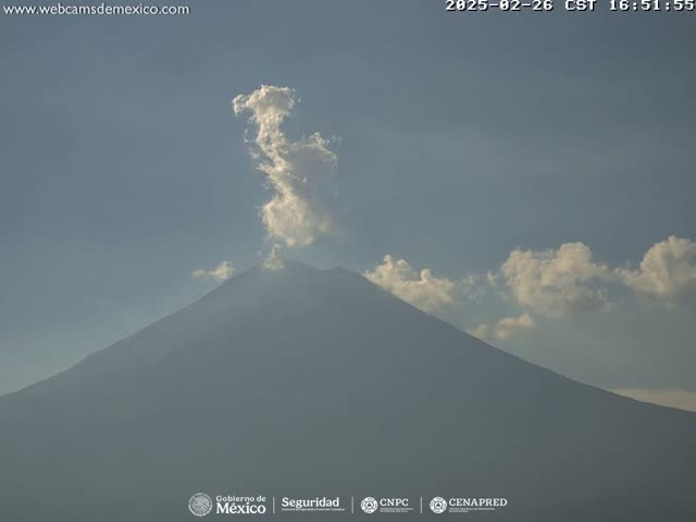 A large, dark mountain is partially obscured by a plume of white smoke and wispy clouds under a mostly clear sky.