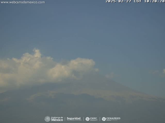 A hazy mountain range is partially obscured by fluffy white clouds under a clear blue sky.