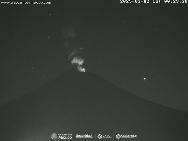 A nighttime, low-light image shows a volcano emitting a plume of smoke or ash under a clear, starry sky.
