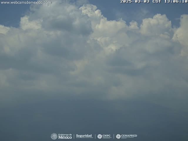 A mostly cloudy sky with fluffy white cumulus clouds against a pale blue background.