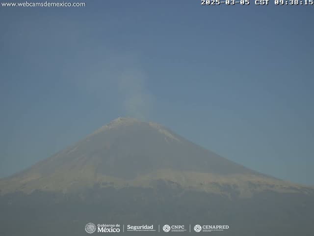 A snow-capped volcano is seen under a clear, hazy sky, emitting a small plume of smoke or steam.