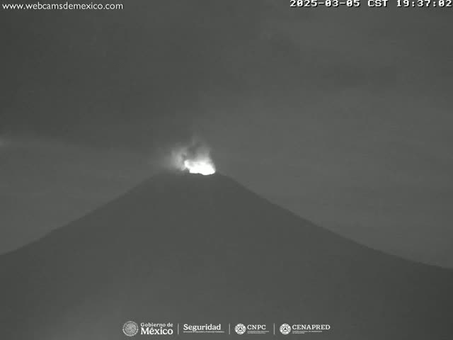A nighttime, low-light image shows a volcano erupting with a bright glow at its peak under a dark, cloudy sky.
