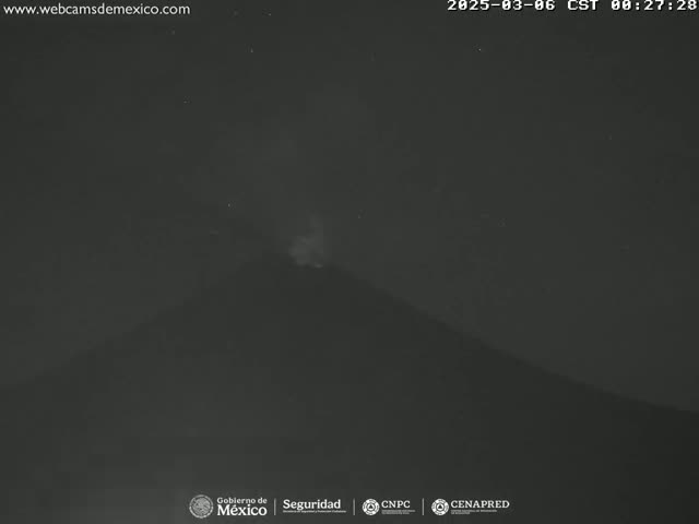 A dark, nighttime image shows a volcano emitting a faint plume of smoke or ash under a clear, starless sky.