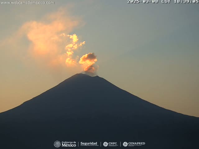 A large, dark volcano is silhouetted against a pale orange and pink sunset sky, with a plume of light-colored smoke or ash emanating from its summit.