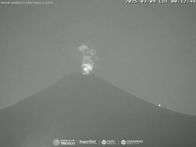 A nighttime, grayscale image shows a volcano emitting a plume of smoke under a dark sky.