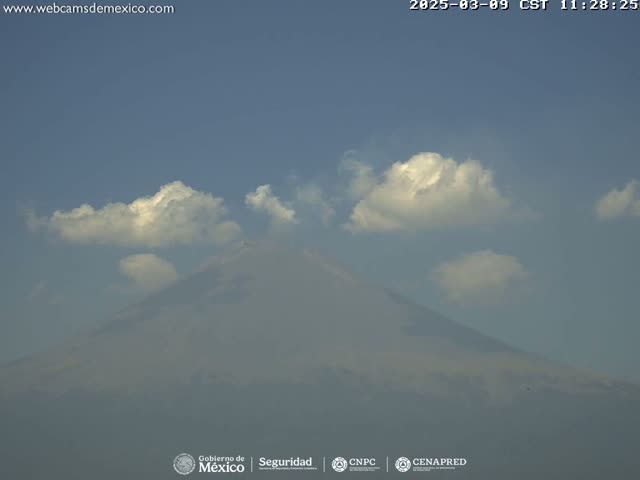 A hazy mountain is partially obscured by fluffy white clouds under a clear blue sky.