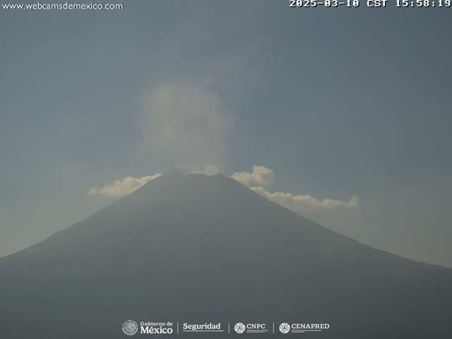 A large, dark volcano is emitting a plume of light gray smoke under a hazy, light-blue sky.